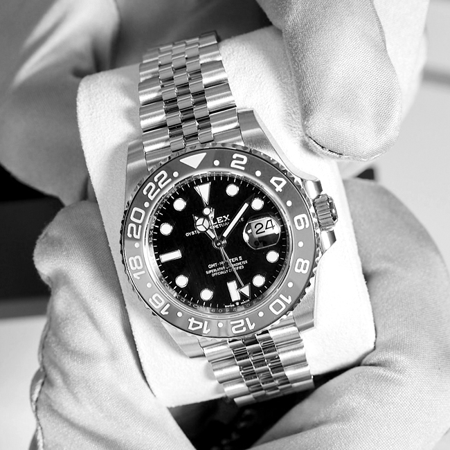 Eine Rolex GMT Uhr, die um ein Uhrenkissen gelegt ist. Dieses wird von Händen in Handschuhen gehalten.